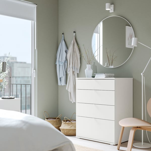 Modern bedroom with green wall, white chest of drawers, round mirror, and wooden chair. Clothes hang on hooks, plants add nature.