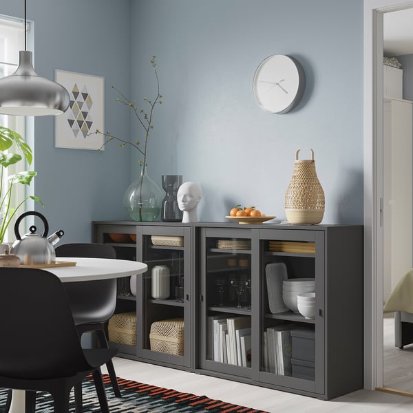 Grey storage unit with glass doors, clock above, holds dishes & books, next to black chair.