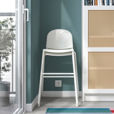 A white junior chair placed between green walls, ideal for kids, fitting between highchairs and regular chairs.