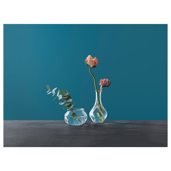 Two glass vases, one with pink flowers, other with greenery, on dark table, teal wall.