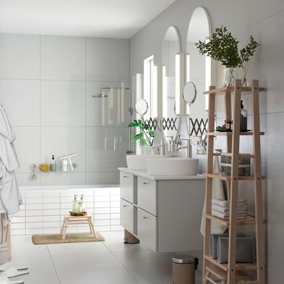 Modern white bathroom with glass shower, dual sinks, mirrors, and a light wood ladder-style shelf stocked with towels and plants.