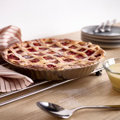 VINTERROCKA pie dish, round, shiny silver, holds a lattice pie. Rest on a wooden table with a spoon and small bowl beside.