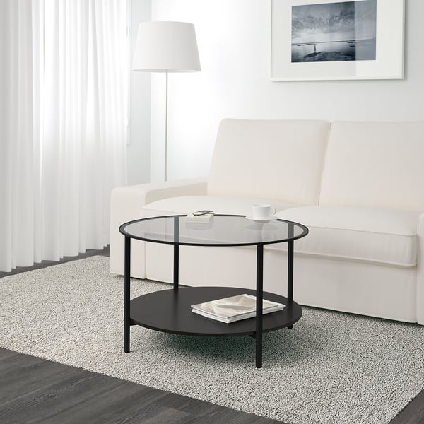 Modern living room with a sleek white couch and a glass-topped VITTSJÖ coffee table holding a cup, saucer, remote, and magazine.