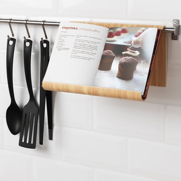 Cookbook holder mounted on kitchen rail, holding open recipe page with chocolate muffins. Three black cooking utensils hang beside it.