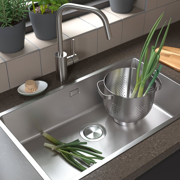 Stainless steel sink with modern tap, colander full of green onions, fresh herbs in pots, scrubber, and cutting board.