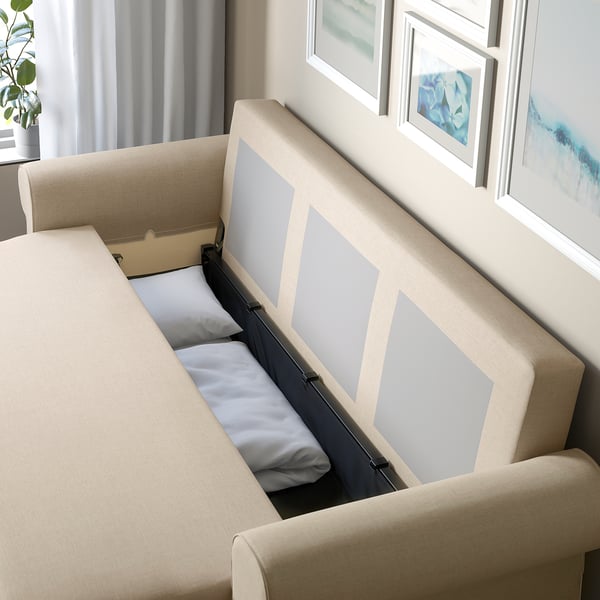 Sofa-bed with storage open, showing bedding inside. Tan upholstery with grey cushions.