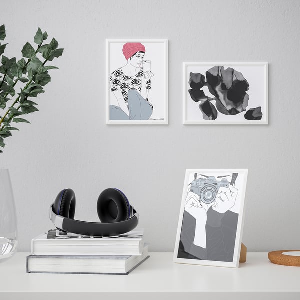 Modern desk setup with art prints, headphones on books, plant in vase. Frames hold changeable pictures, one stands.