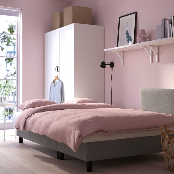 Jaukus miegamasis su rožinėmis sienomis, patalyne ir šviesa. Balta spinta, sieninė lentyna su dekoru ir sieninis šviestuvas.