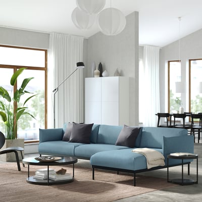 Moderni svetainė su ÄPPLARYD mėlyna sofa, stilingomis pagalvėlėmis, aptakiais kavos staliukais, dideliais langais ir aukštais kambariniais augalais.