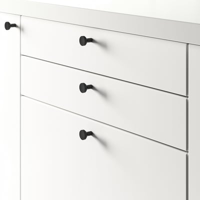 Balta komoda su keturiais stalčiais, kurių kiekvienas pritvirtintas juodomis rankenėlėmis, tinka įvairaus storio durims.