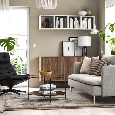 Moderni svetainė su BANKKONTOR kilimu, pilka sofa, žurnaliniu staliuku, juoda kėde, medžio saugykla ir augalais. Kilimėlių dydis papildo baldus.