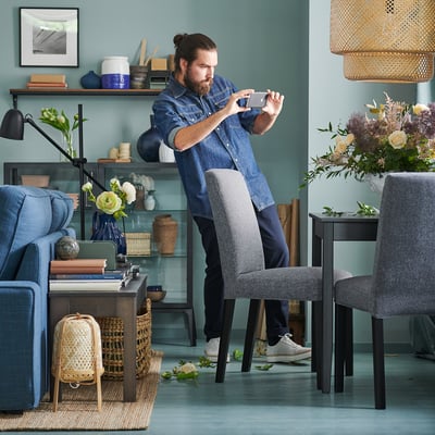 Vyras džinsiniame audinyje fotografuojasi mėlyname kambaryje prie kėdės ir sofos.