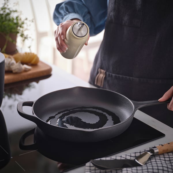 Asmuo pagardina keptuvę aliejumi iš butelio. Keptuvė ant viryklės, peilis netoliese. Stalviršis turi ingredientų, augalų.