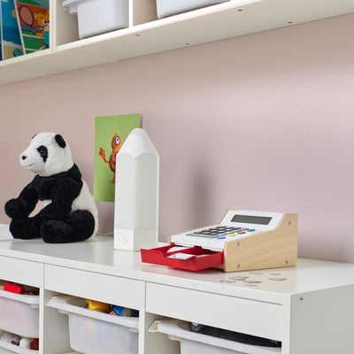 Baltas žaislinis kasos aparatas su skaičiuokle ant stalo, žaidybiniai pinigai, rausva siena.