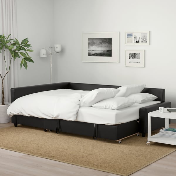 Juoda sofa-lova FRIHETEN su baltomis pagalvėmis minimalistiniame kambaryje. Nuotraukos, augalas, šoninis stalas su žurnalais. Šviesus kilimas, medinės grindys, pilkos užuolaidos.