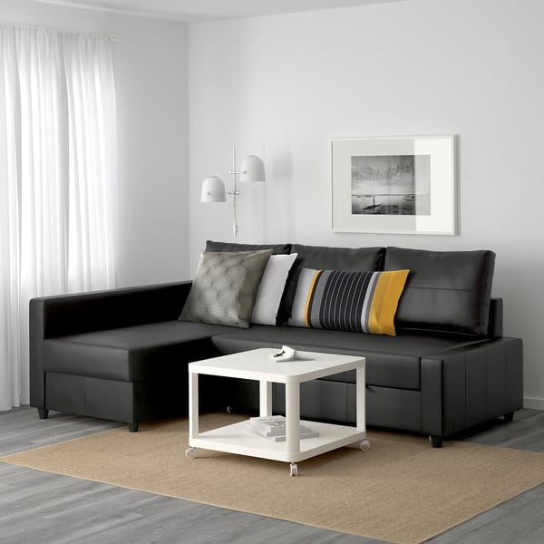 Moderni minimalistinė svetainė su juoda FRIHETEN sekcijine sofa, spalvingomis pagalvėmis, baltu kavos staliuku, smėlio spalvos kilimu, grindų lempa ir sienų menu.