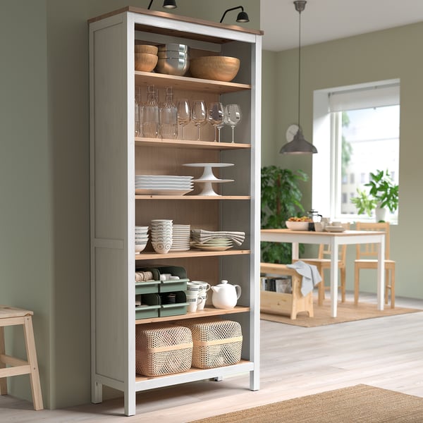 HEMNES knygų spinta su reguliuojamomis lentynomis, talpinančiomis taures, dubenėlius ir puodelius. Šviesiai ruda, šalia valgomojo komplekto, su juoda lempa viršuje.