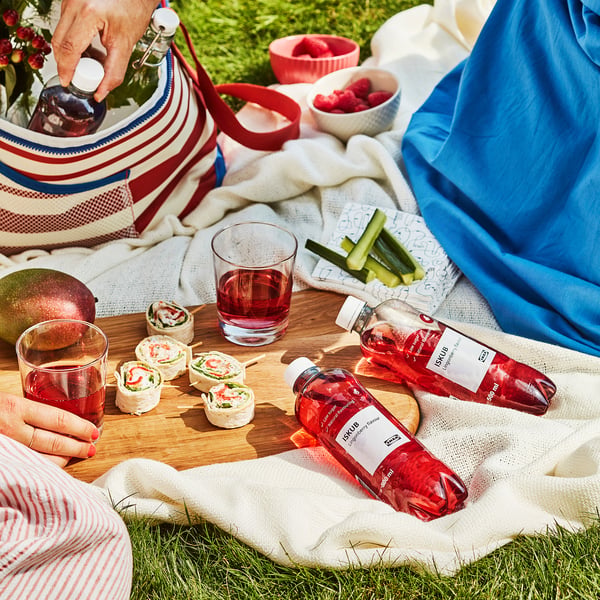 Pikniko scena su ISKUB gėrimais, sumuštinių suktinukais, vaisiais ir užkandžiais, išdėstytais ant antklodės. Kažkas pila ISKUB gėrimą į stiklinę.