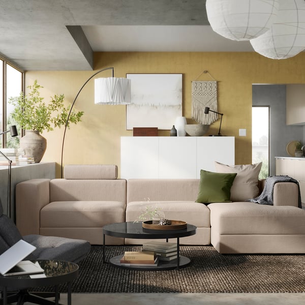 Moderni svetainė su smėlio spalvos JÄTTEBO sofa, geltona siena, pilko ploto kilimėliu ir kabančiais žibintais.