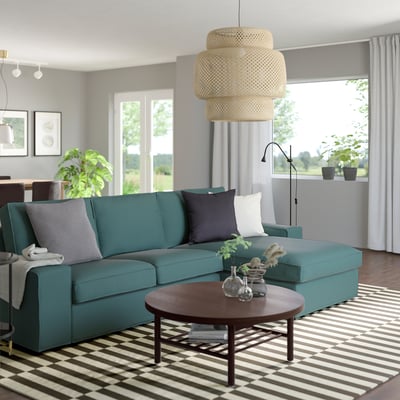 Teal KIVIK sekcijinė sofa su dryžuotu kilimu, apvaliu kavos staliuku ir pakabinamu žibintu. Atvira erdvė su baltomis užuolaidomis ir augalais.