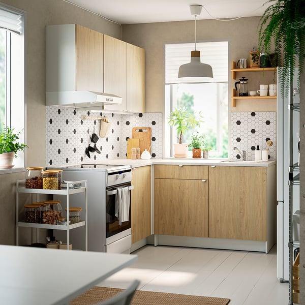 Šiuolaikinė l-virtuvė: šviesios medinės spintelės, marmuro skaitikliai, juodas backsplash, aukšta viryklė, metalinis vežimėlis, pakabukai, dideli langai, natūrali šviesa.