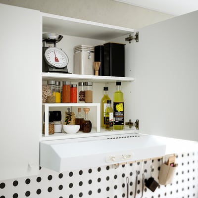 Atidarykite virtuvės spintelę, kurioje rodomi įvairūs stiklainiai, buteliai ir svarstyklės. Lentyna reguliuojama. Juoda polka dot backsplash.