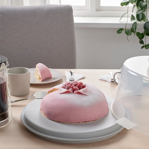 Tortas laikiklyje su dangčiu ir rankena, parodytas ant stalo su lėkšte ir puodeliais.