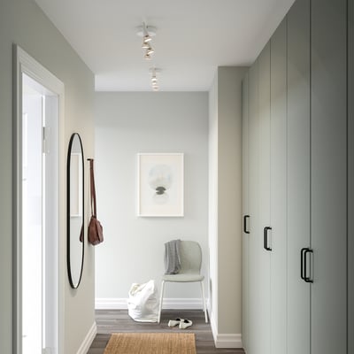 Modernus įėjimas su lubų šviesa, šviečiančia pilka kėde ir rudu krepšiu ant sienos kablio. Aukšta spinta dešinėje, baltos durys kairėje.