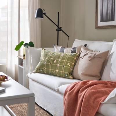 Svetainė su balta sofa, KUSTGRAN pagalve, dekoratyvinėmis pagalvėmis, antklode, lempa ir augalu.