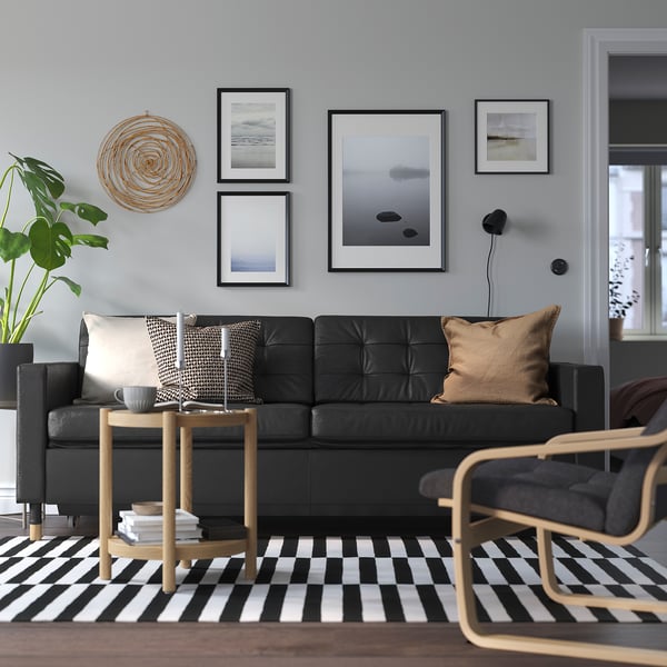 Juoda odinė sofa su pagalvėlėmis ir kavos staliuku, languotas kilimas, sienų menas, jauki atmosfera.