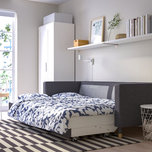 Modernus miegamasis su konvertuojama sofa-lova ant ratų, baltos lentynos su knygomis ir dekoru, didelė drabužių spinta ir dryžuotas kilimas natūralioje šviesoje.