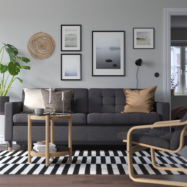 Jauki pilka sofa LANDSKRONA su medinėmis kojomis. Jos didelės, su pagalvėlėmis ir kavos staliuku šalia. Meno kūriniai ir augalų dekoras modernioje svetainėje.