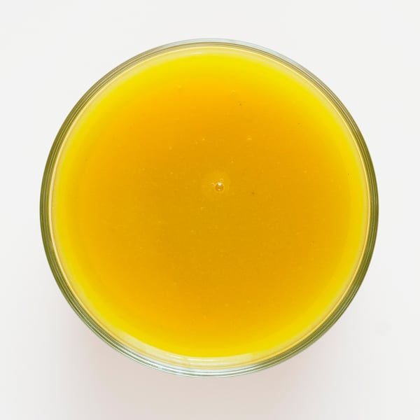 Skaidrus, apvalus stiklinis dubuo, užpildytas ryškiu oranžinės geltonos spalvos kokteiliu, pagamintu iš mango, moliūgo ir obuolio, demonstruojantis sklandžią tekstūrą.