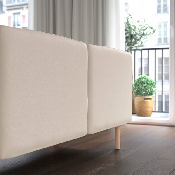 Smėlio spalvos LILLEHEM sofa su medinėmis kojomis prie lango. Matomi augalai ir miesto vaizdas.