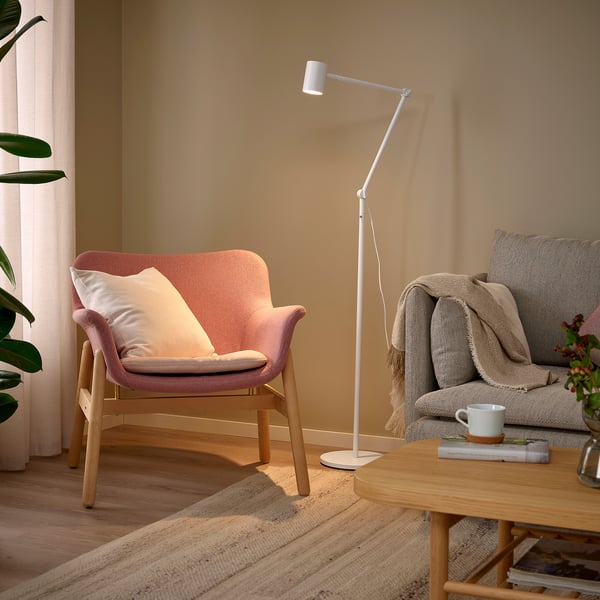 Jauki svetainė su NYMÅNE grindų lempa, šviesiu medžiu ir neutraliais tonais. Rausva kėdė, pilka sofa, reguliuojamas šviestuvas, medinis kavos staliukas ir kilimėlis.