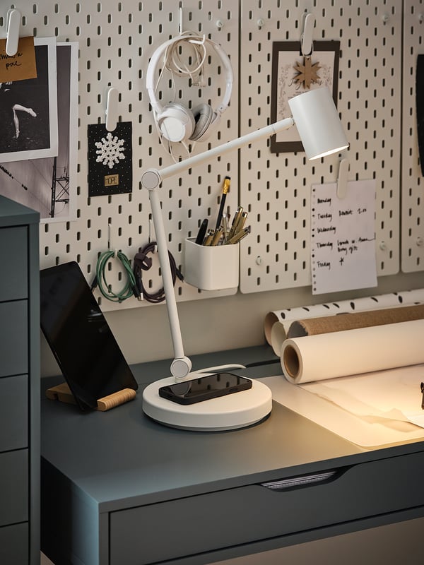 Moderni darbo vieta su NYMÅNE lempa, telefonu, rašiklio laikikliu, popieriumi ir lenta su užrašais ir ausinėmis.