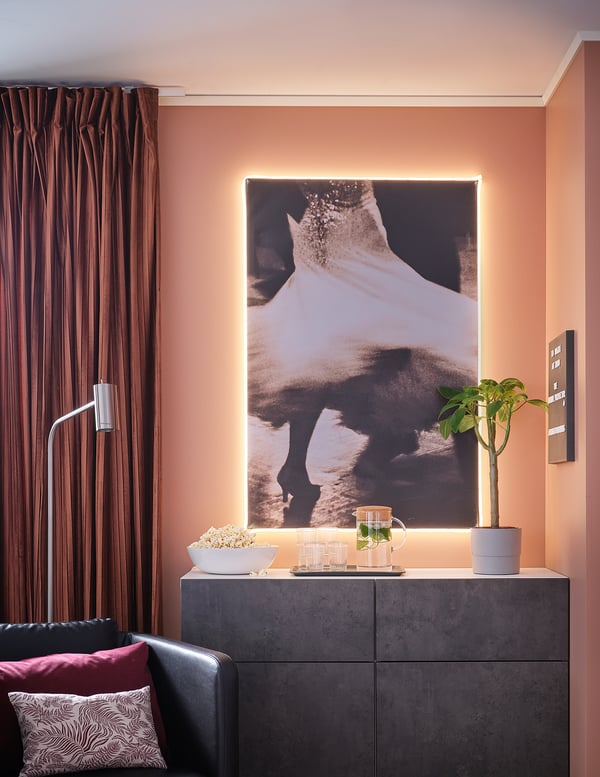 Moderni svetainė su persikų sienomis, juoda spinta, abstrakčiais meno kūriniais, juoda sofa, pagalvės, spragėsiai, augalai, rudos užuolaidos, led žibintai.