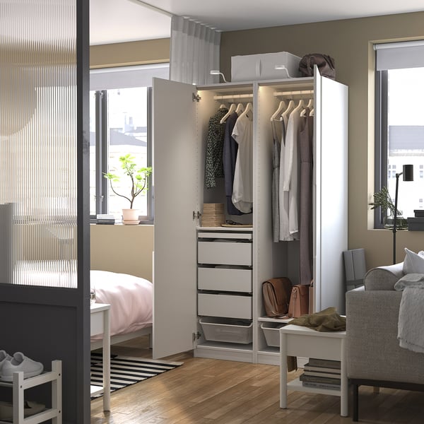 Modernus miegamasis su balta PAX spinta, atviras rodyti pakabas, stalčius ir daiktadėžes. Kambaryje yra lova, langai, augalas ir fotelis.