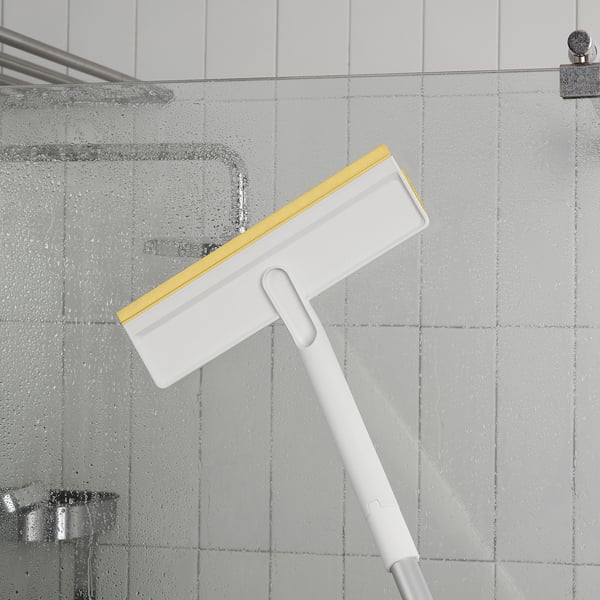 Balta teleskopinė valymo rankena su geltonos ir baltos spalvos valytuvu, valanti dušo sienelę vandens lašeliais.