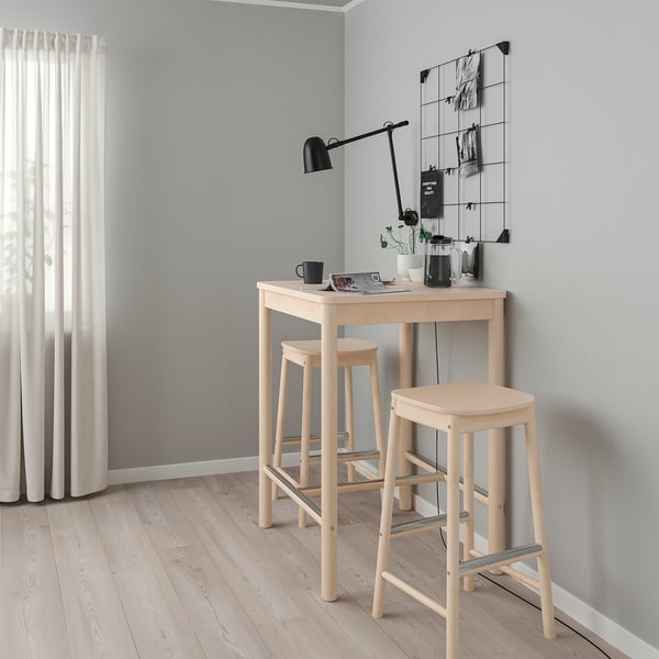 Nedidelis šviesus medinis RÖNNINGE baro staliukas su taburetėmis minimalistiniame kambario kampe, balta užuolaida, pilka siena, juodas šviestuvas.