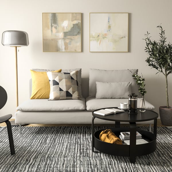 Moderni svetainė su sofa SÖDERHAMN, balta su raštuotomis pagalvėmis, apvalus juodas kavos staliukas, grindų lempa ir abstraktus menas ant sienų.