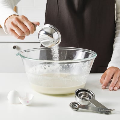 Asmuo prideda ingredientų į maišymo dubenį, naudodamas matavimo puodelius. Puodeliai dedami ant prekystalio, paruošti tiksliems matavimams.