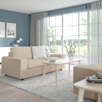 Moderni svetainė su smėlio spalvos sofa VIMLE, šviesiu mediniu kavos staliuku, dviem šoniniais staliukais, kilimėliu, lempa, vaza ir užuolaidomis. Sofa prie didelių langų.