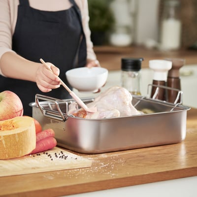 Человек, готовящий мясо в нержавеющей стальной форме для жарки с решёткой, окружённый овощами и специями.