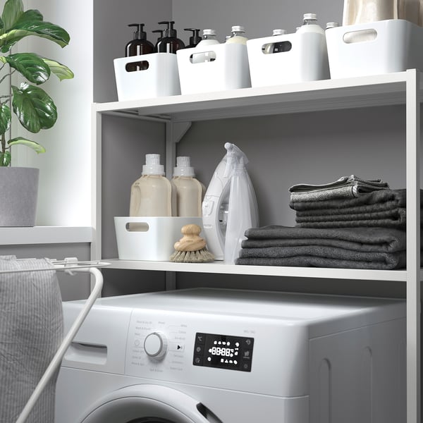 Белая полка в прачечной над стиральной машиной, в которой хранятся полотенца, моющие средства и утюг.