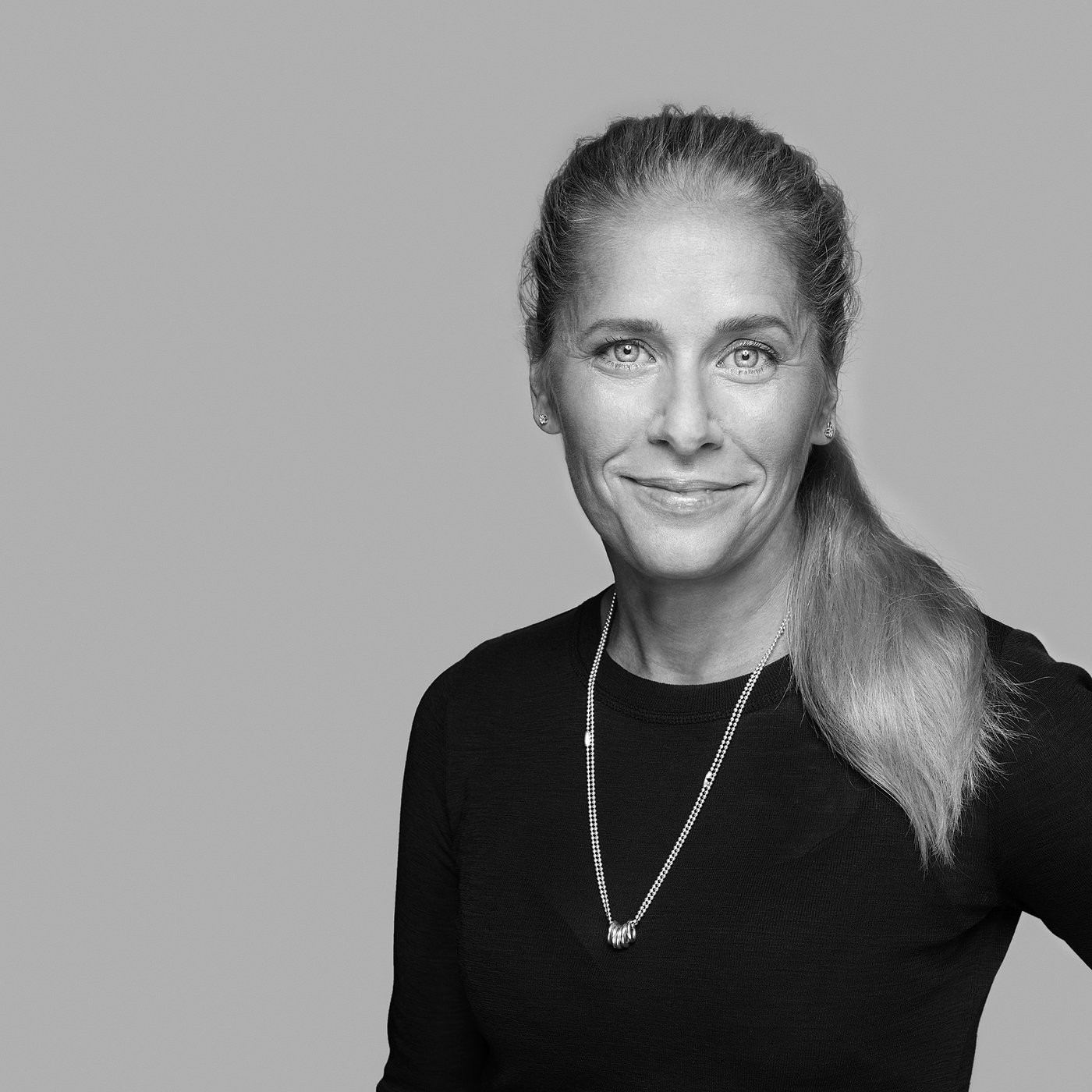 Черно-белый портрет улыбающейся Sarah Fager в ожерелье и тёмном топе.