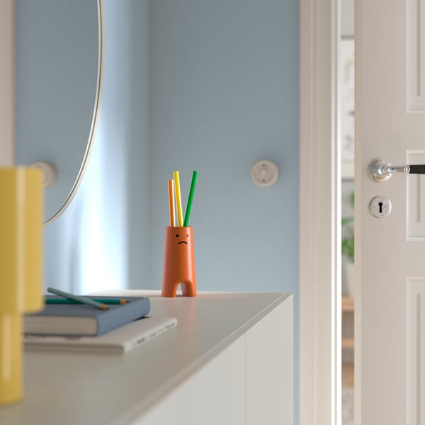 Оранжевая ваза с грустным лицом, держащая карандаши рядом с блокнотами и линейкой.