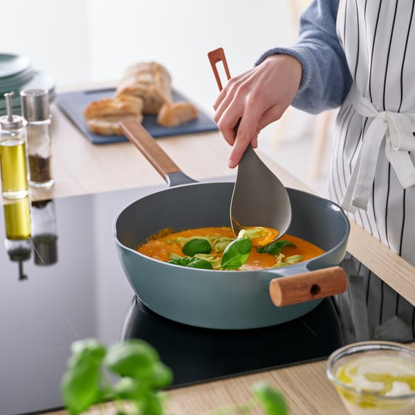 Женщина готовит разноцветной KNORRHANE глубокой ложкой, соскребая тушенку со сковороды. Ложка стоит на плите вместе с другими кухонными принадлежностями.
