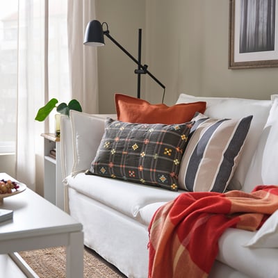 Современное жилое пространство с большой серой подушкой KUSTGRAN, оранжевыми подушками и красным одеялом на белом диване. Черная лампа и растение рядом.