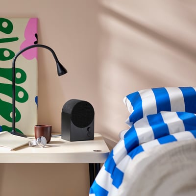 Черная bluetooth-колонка на светлом столе, красочные произведения искусства рядом, яркое постельное белье.